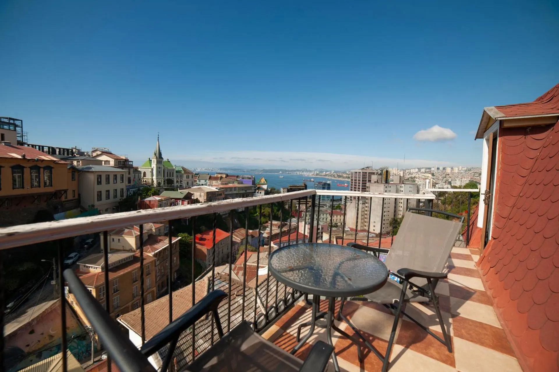Una terraza con mesa de vidrio y sillas, mirando a los techos del cerro, una iglesia con campanario y la bahía.
